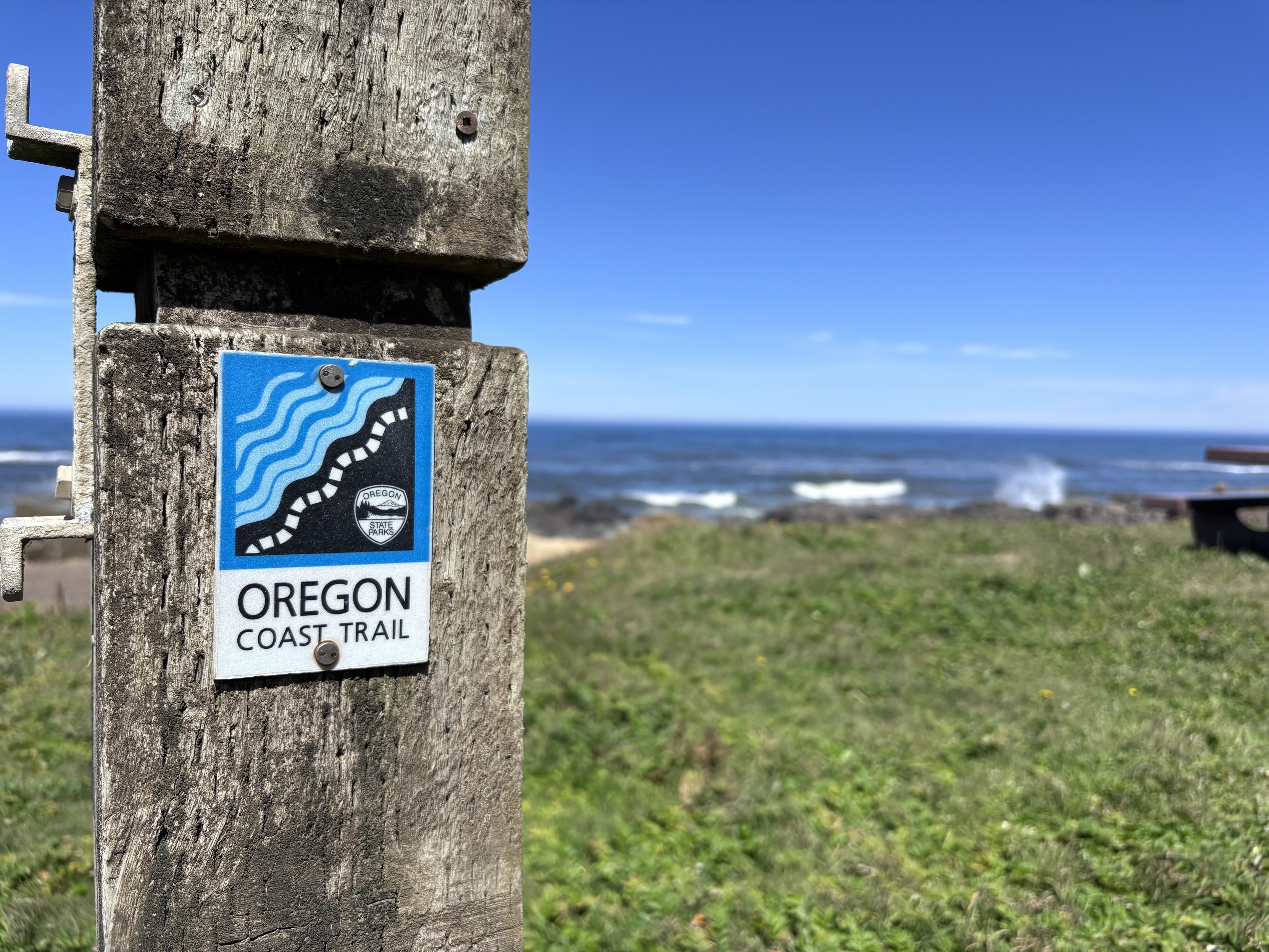 A weathered wooden post displays a sign for the Oregon Coast Trail, with a blue sky and ocean in the background. Green grass and a hint of rocky shoreline are visible.
