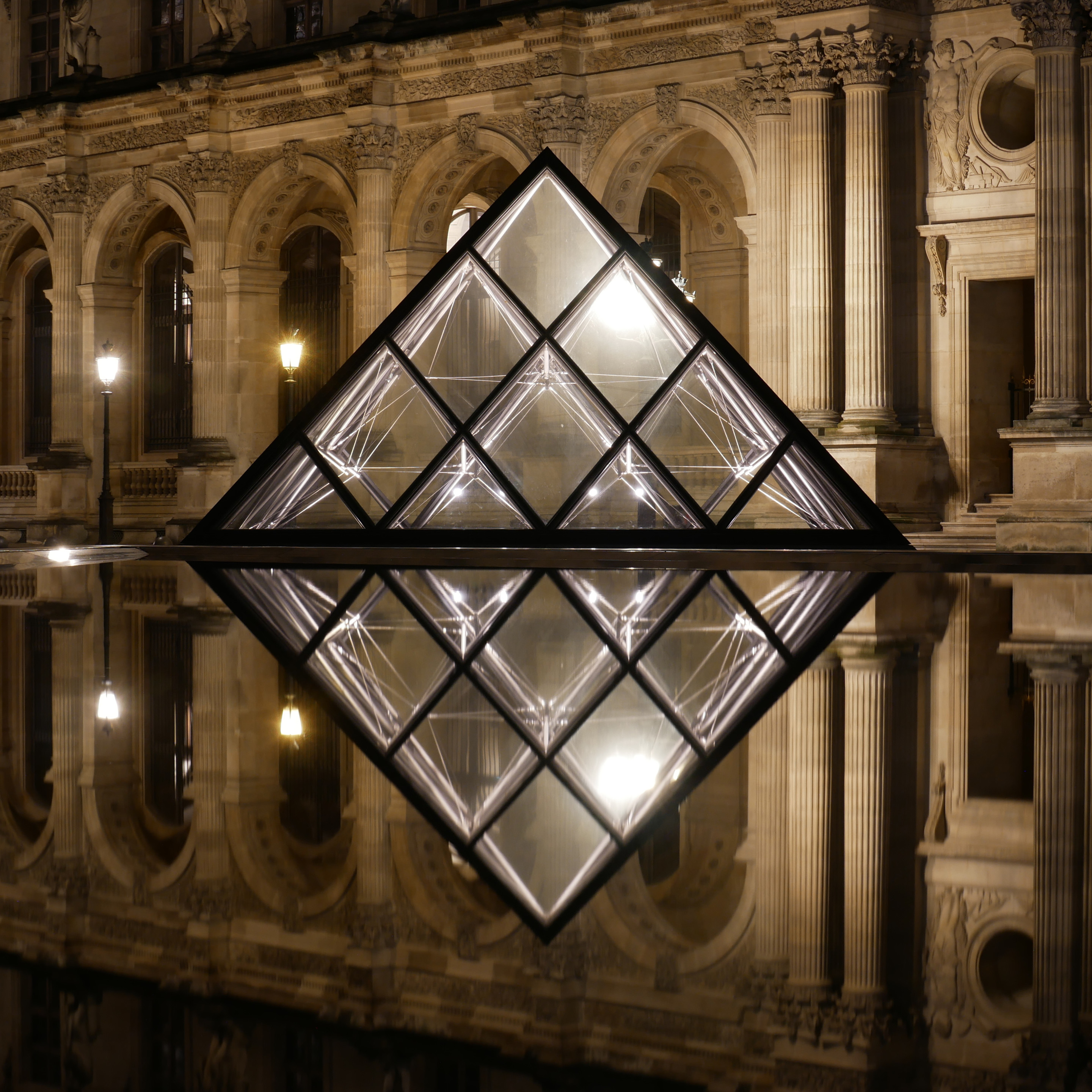 Vorderansicht einer der Pyramiden im Hof des Louvre in Paris, einer Konstruktion aus Glas und Stahl. Das Foto wurde bei Nacht aufgenommen, sodass sich die beleuchtete Pyramide im Wasser des Brunnens davor spiegelt, und sich aus der dreieckigen Form mit Spiegelung ein Quadrat ergibt.
Im Hintergrund die Säulengänge des Louvre mit altmodischen Straßenlaternen. Vorderansicht einer der Pyramiden im Hof des Louvre in Paris, einer Konstruktion aus Glas und Stahl. Das Foto wurde bei Nacht aufgenommen, sodass sich die beleuchtete Pyramide im Wasser des Brunnens davor spiegelt, und sich aus der dreieckigen Form mit Spiegelung ein Quadrat ergibt.
Im Hintergrund die Säulengänge des Louvre mit altmodischen Straßenlaternen.