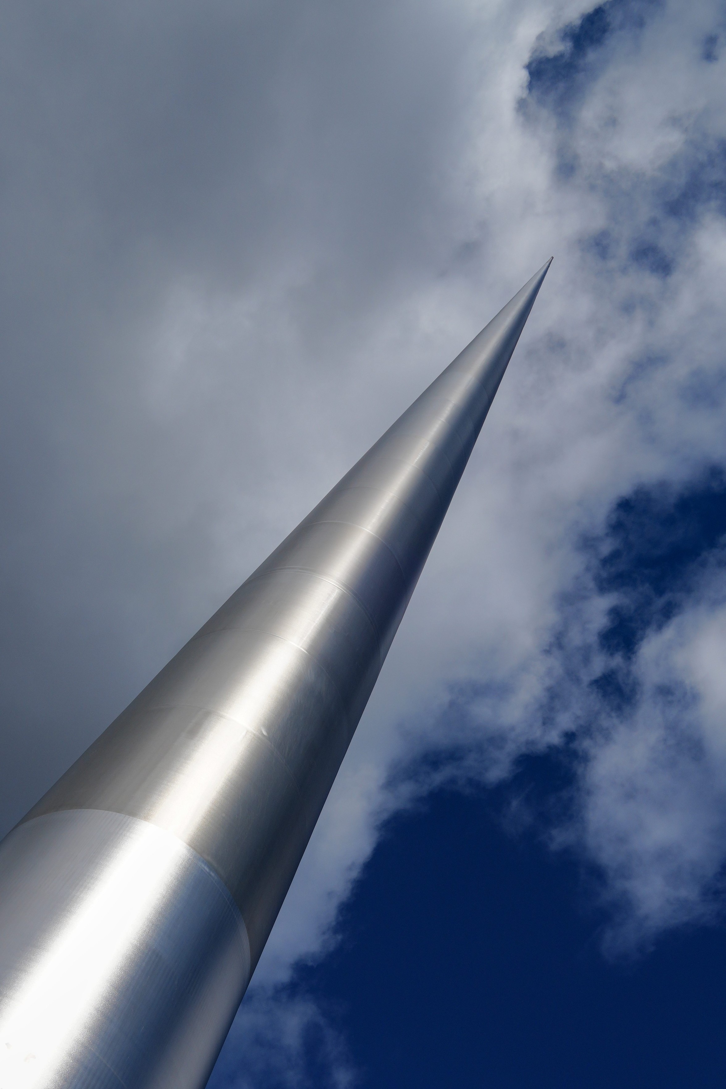 Ein Foto im Hochformat zeigt die Spitze des Monuments "The Spire" in Dublin, eine stählerne, riesige Nadel, die 120 Meter hoch ist. Das Monument zeigt diagonal von links unten nach rechts oben. Im Hintergrund sieht man Wolken vor dunkelblauem Himmel.