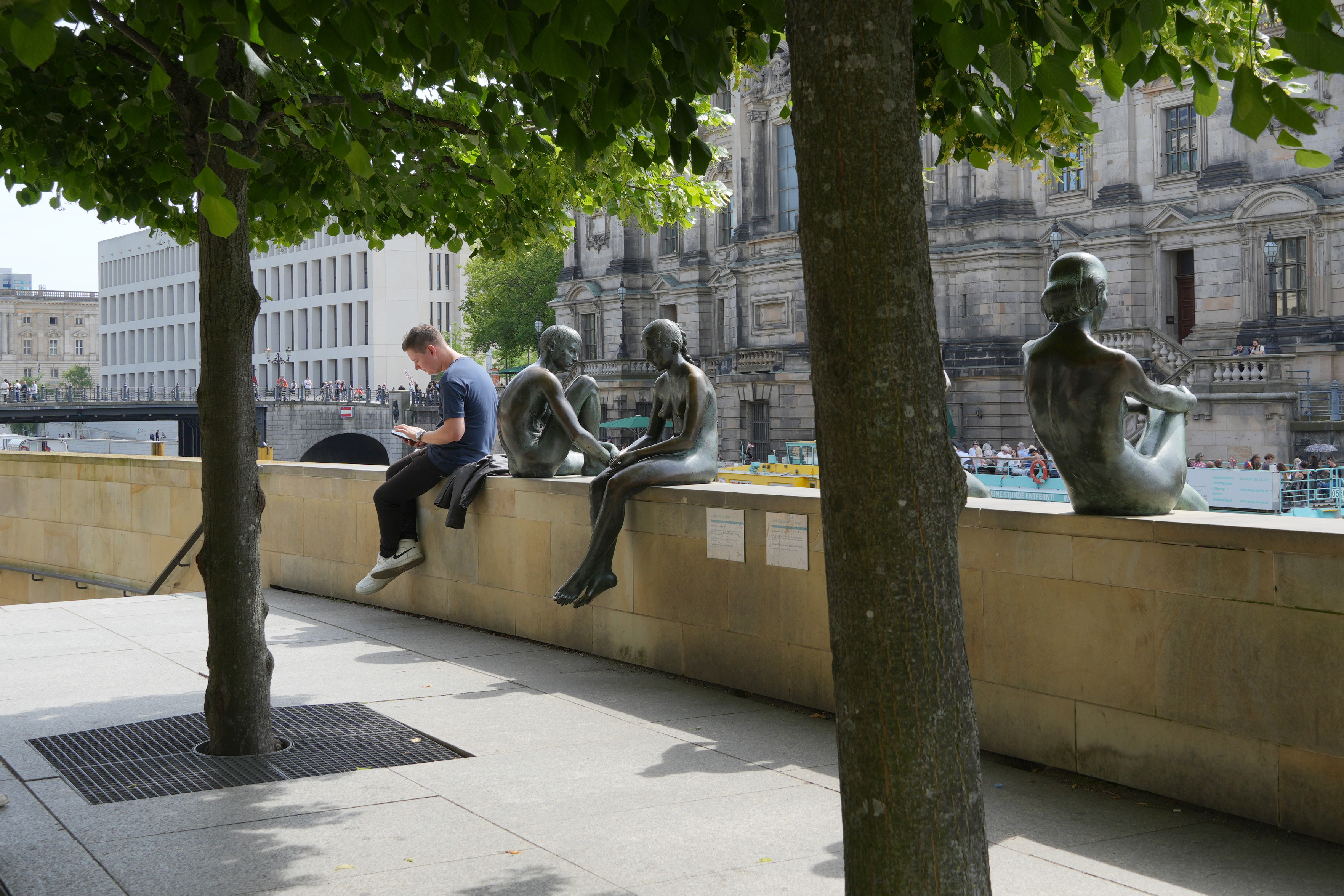 Zwischen zwei Bäumen hindurch sieht man eine schräg durchs Bild verlaufende Mauer, auf der Bronzefiguren von Menschen sitzen, sowie ein echter Mann, der auf sein Handy schaut. Der Mann sticht heraus, da er im Gegensatz zu den Figuren als einziger nicht bronzefarben und oxidiert ist, sich aber sonst perfekt in die Reihe der sitzenden einfügt. Im Hintergrund sieht man Häuserfassaden der Berliner Innenstadt.