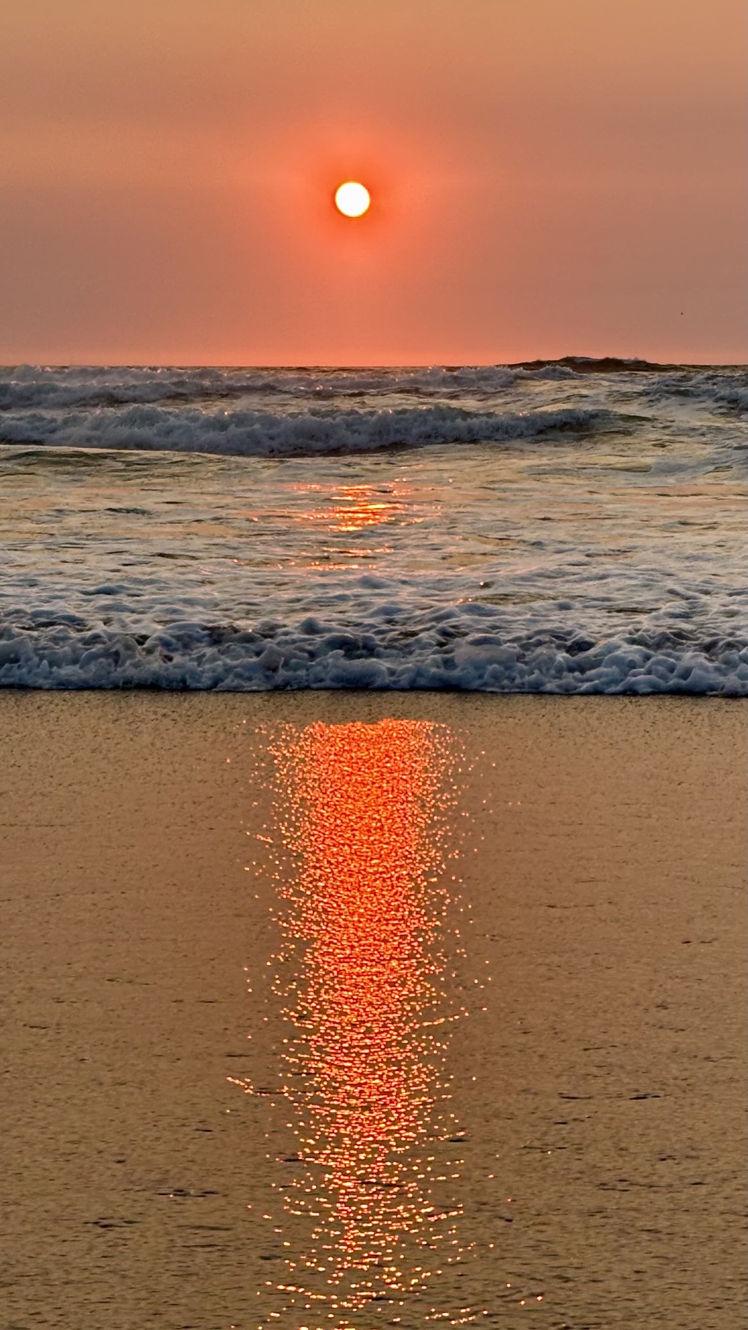 A serene beach scene at sunset, featuring a vibrant orange sun dipping below the horizon. The water reflects the warm hues, creating a shimmering path of light on the sand. Gentle waves roll in, enhancing the peaceful atmosphere.