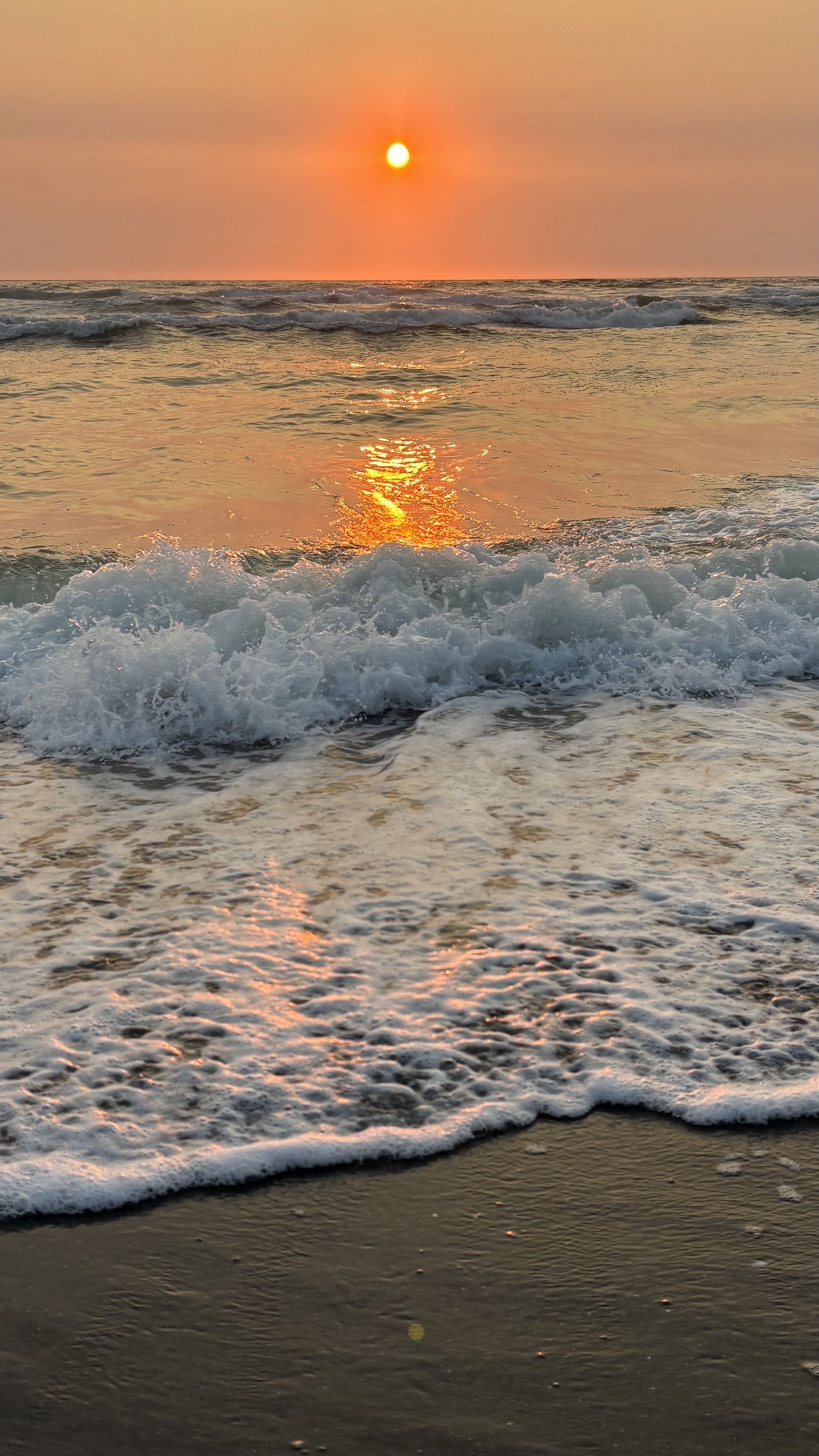 A sunset over the ocean is depicted, with vibrant orange and yellow hues filling the sky. Waves gently crash onto the shore, reflecting the warm colors of the sunset. The scene evokes a sense of tranquility and natural beauty.