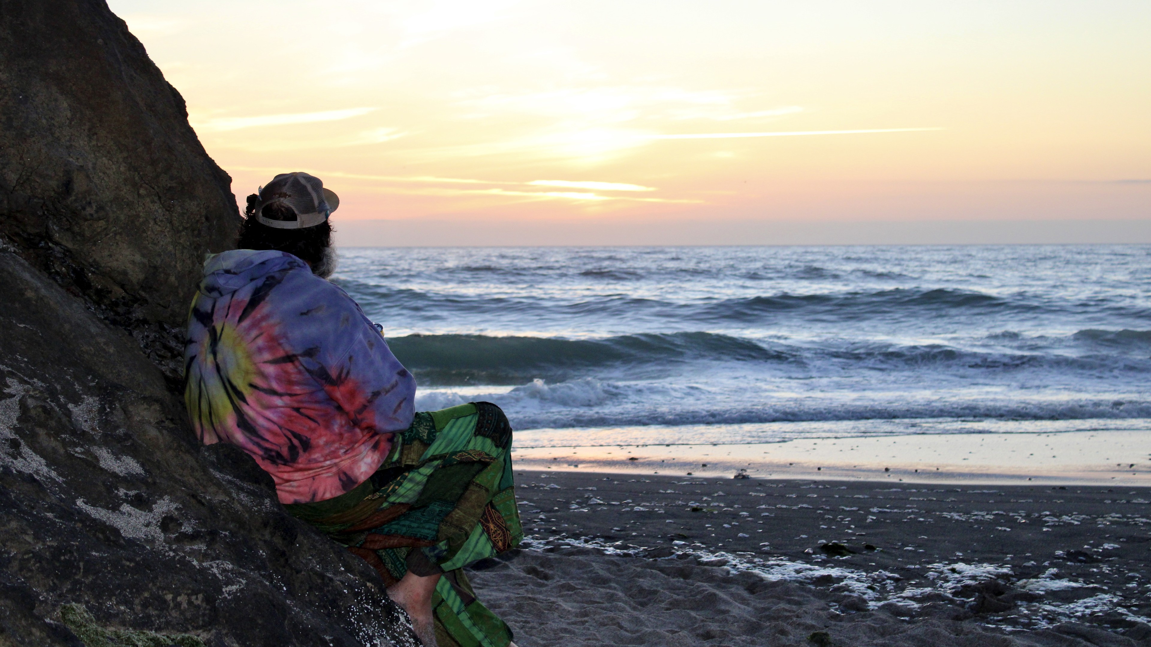 Person sitting on a rocky beach at sunset, wearing a colorful tie-dye shirt, facing the ocean.