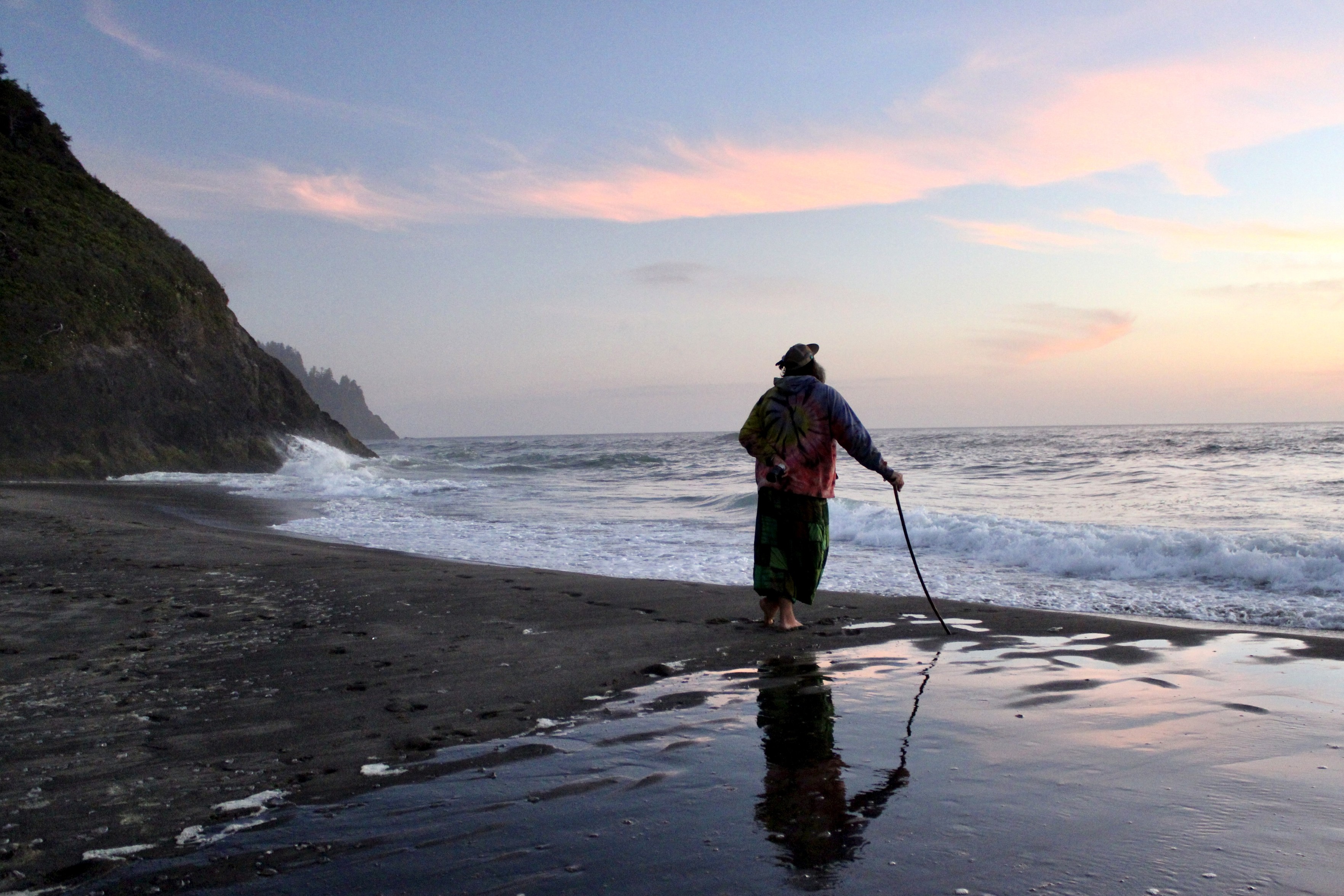 Person standing on a dark beach at sunset, holding a walking stick beside the ocean waves.