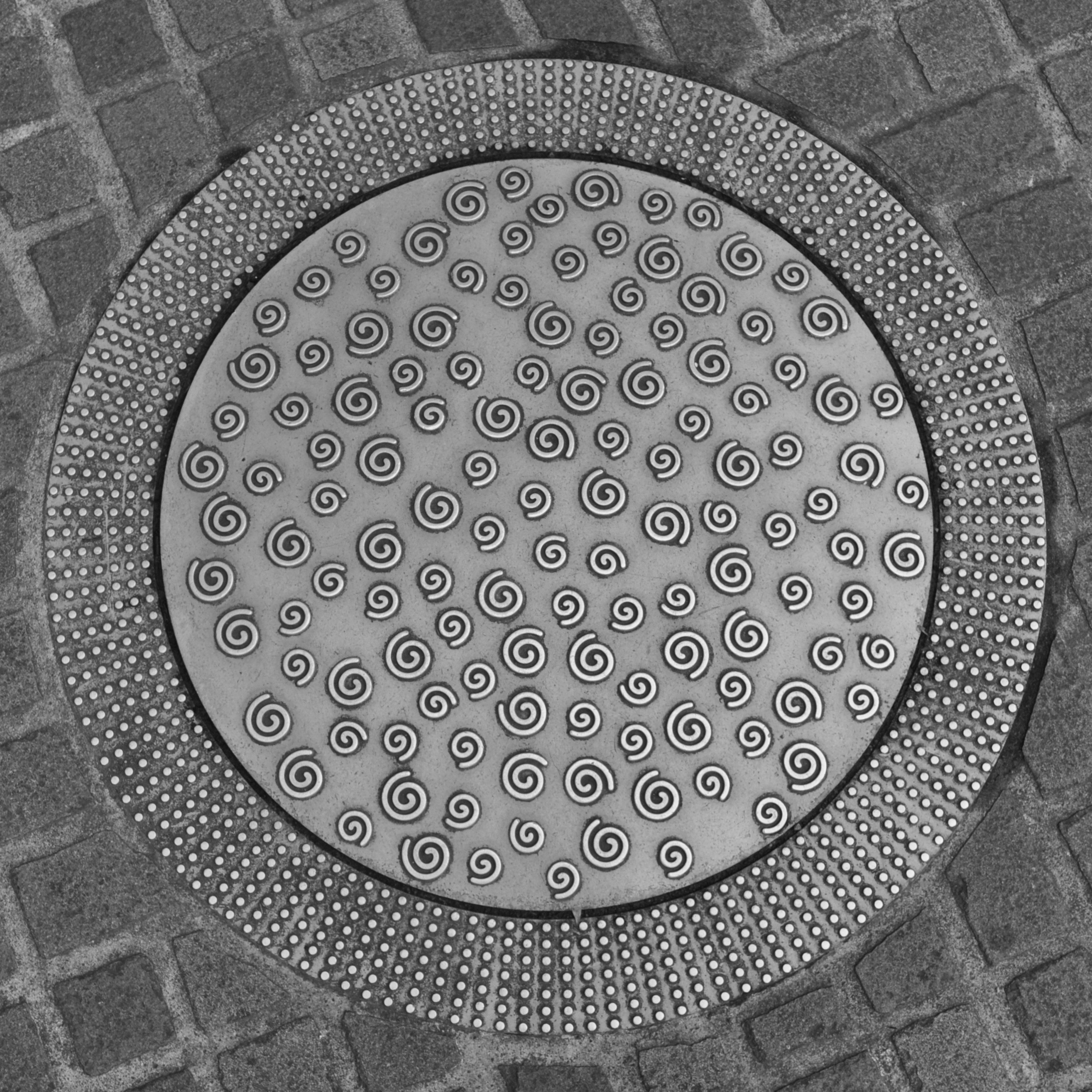 Ein in Kopfsteinpflaster eingelassener, silberfarbener Kanaldeckel mit ungewöhnlicher Dekoration: der Deckel selbst ist glatt poliert und mit unzähligen kleinen Spiralen verziert. Der Rahmen des Deckels ist in mit runden Noppen in konzentrischen Kreisen überzogen.