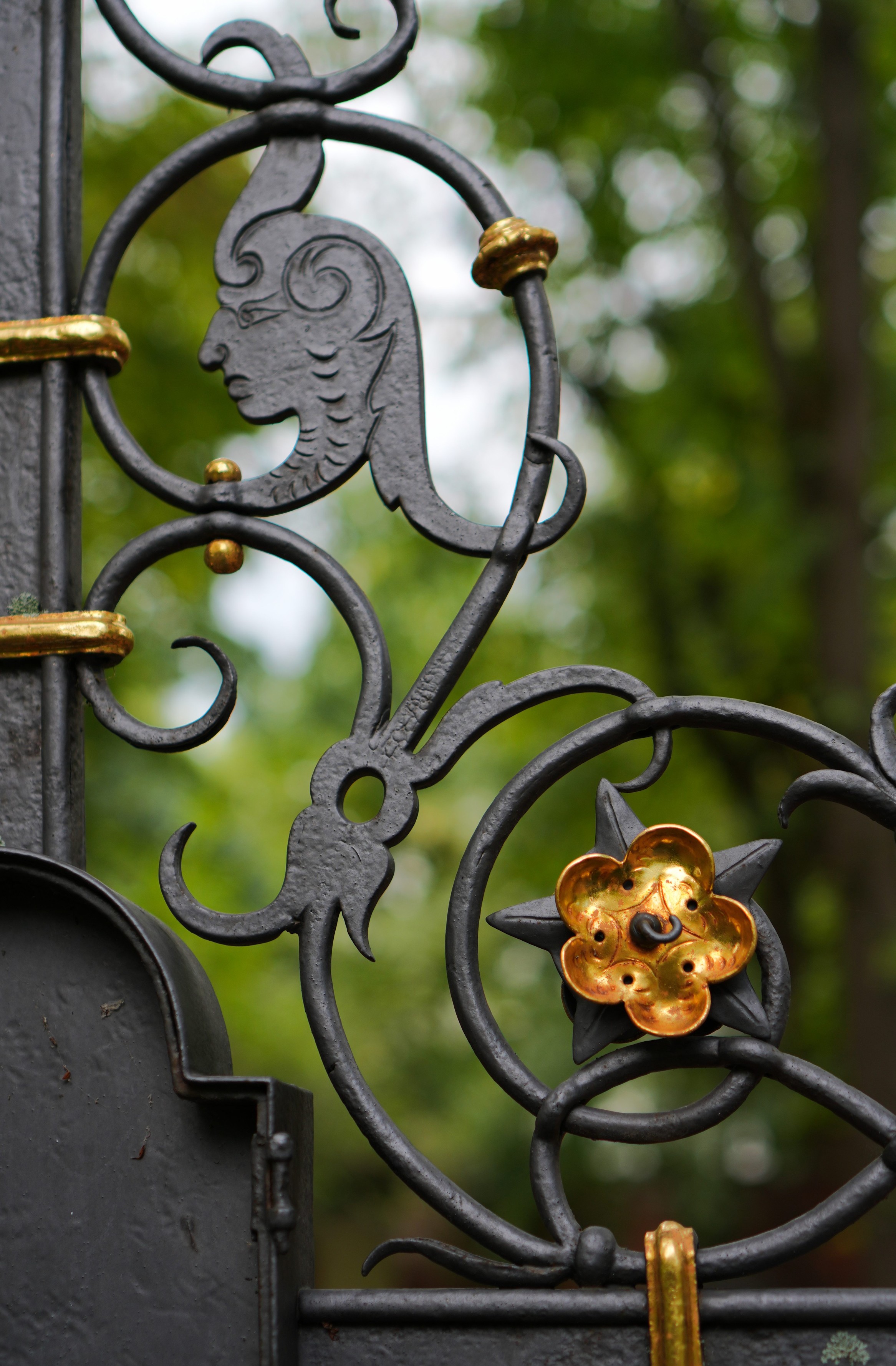A close-up photo of metalworking details on a grave. Filigree floral elements in black are combined with a golden and well-polished blossom piece as well as a relief of the head of some kind of fairy tail figure. The background is all blurred, but a tree and foliage can be made out.