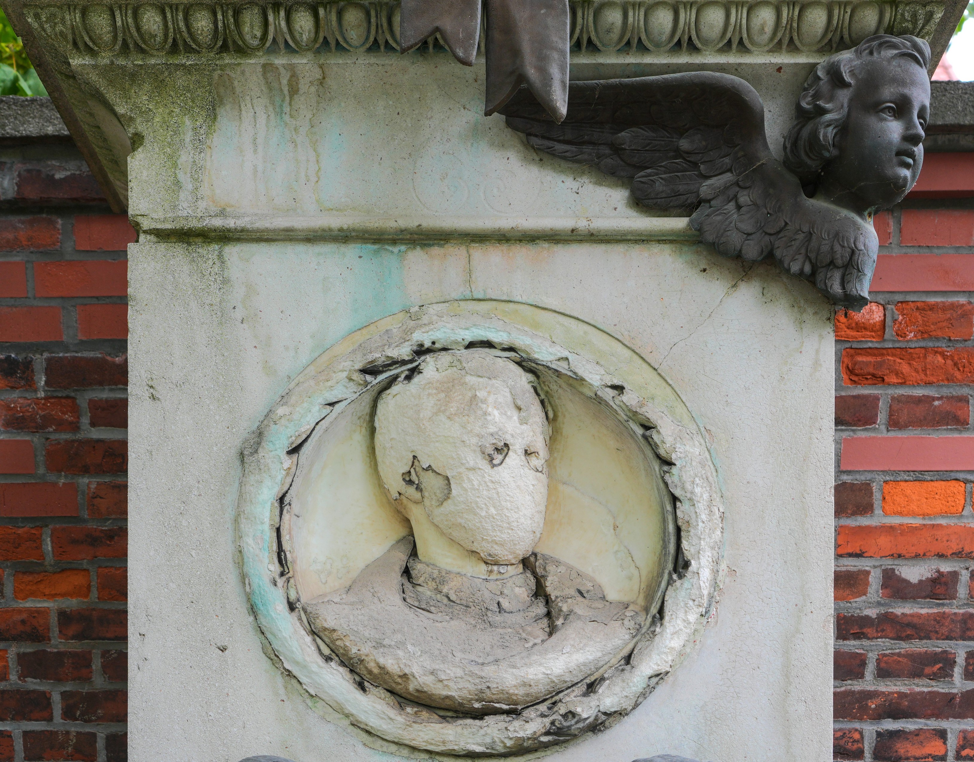 A large pillar that probably used to a memorial to a long-dead man, who used to be displayed in a relief. Most of the paint and plaster of it has come off though, so all you can make out is a head with two eye sockets, no other facial features, but a collar. The pillar is adorned with a bust of an angel, possibly bronze, darkened by time and weather, but otherwise in good shape.