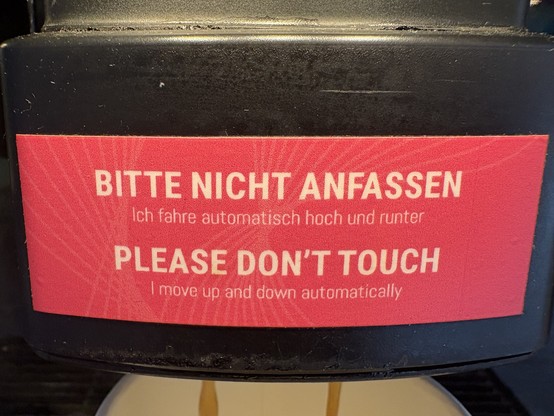 Ein rotes Schild mit der Aufschrift „BITTE NICHT ANFASSEN“, „Ich fahre automatisch hoch und runter“ und „PLEASE DON'T TOUCH“, „I move up and down automatically“.